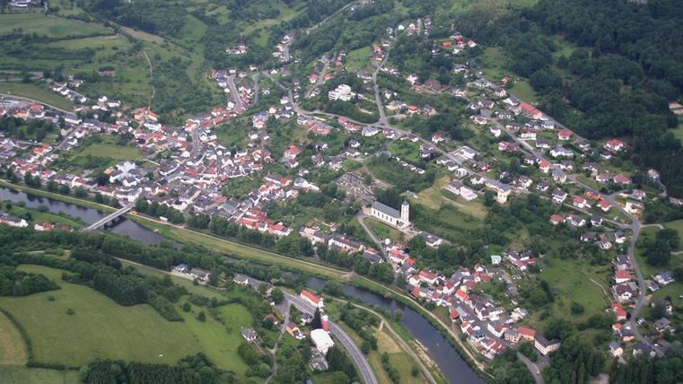 Eine Luftaufnahme eines kleinen Dorfes mit vielen Häusern und einem Fluss. Umgeben von grünen Wiesen und Hügeln.