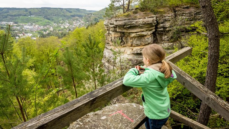 Ein Mädchen steht an einem Geländer und blickt auf eine grüne Landschaft mit Felsen und Bäumen. Im Hintergrund sind hügelige Landschaften zu sehen.