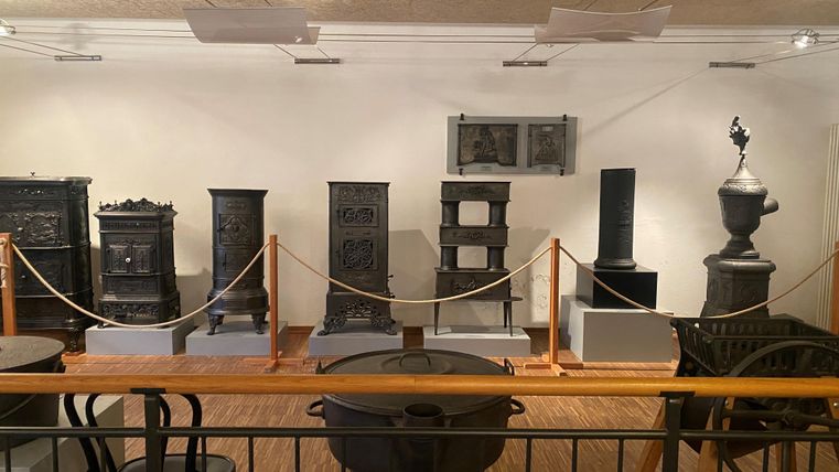 Eine Ausstellung mit antiken Öfen und Heizkörpern in einer Museumsdarbietung. Die Objekte sind auf Podesten platziert und durch eine Absperrung gesichert.