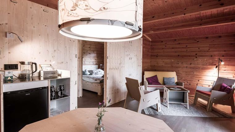 Ein gemütlicher Raum mit Holzkonzept, einer Esstischmitte und einem großen Licht. Auf der rechten Seite befinden sich bequeme Sitzgelegenheiten und Zugang zu einem Schlafzimmer.