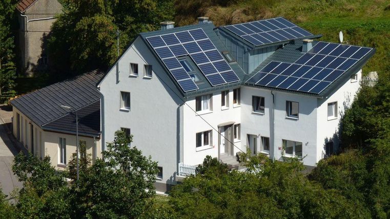 Ein modernes Haus mit Solarpanelen auf dem Dach. Es ist von Bäumen und Grünflächen umgeben.