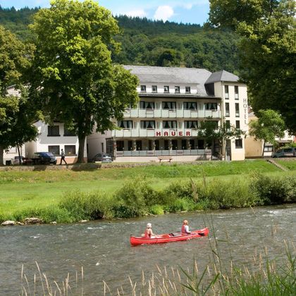 Ein schönes Hotel am Fluss mit grünen Bäumen und einem klaren Himmel. Zwei Personen paddeln im roten Kanu auf dem Wasser.