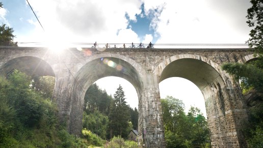 Radfahrer überqueren das Reichensteiner Viadukt der Vennbahn. Die Sonne scheint durch die Wolken, während das Viadukt über eine grüne Landschaft führt., © vennbahn.eu