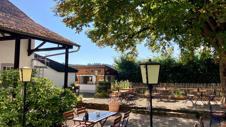 Un espace extérieur accueillant avec des tables et des chaises sous un arbre. Le ciel est clair et l'environnement semble calme et amical.