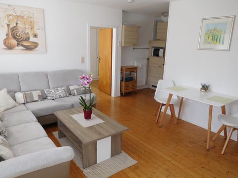 Ein helles Wohnzimmer mit einer gemütlichen Couch, einem Tisch und einem kleinen Esstisch. Eine Orchidee in einem Topf sorgt für einen frischen Akzent im Raum.