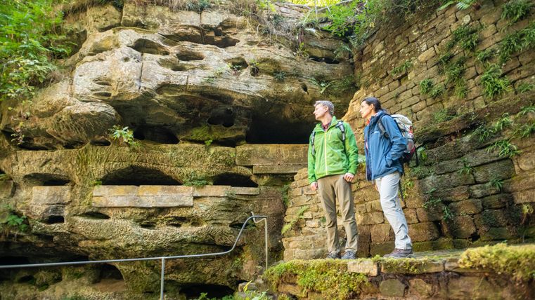Zwei Wanderer stehen auf einem Felsen und betrachten eine Felswand mit Löchern. Umgeben von üppigem grünem Bewuchs erleben sie die natürliche Schönheit der Umgebung.