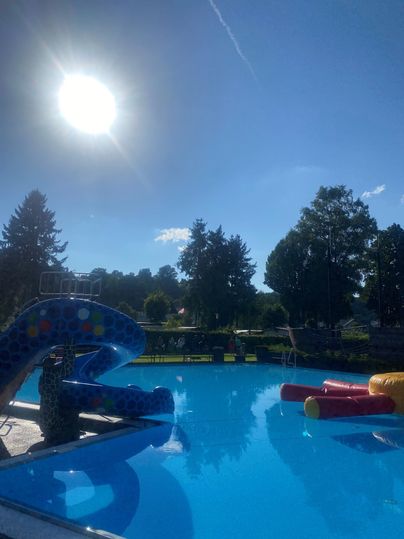 Ein Schwimmbad mit einer Rutsche und bunten Wasserattraktionen. Der Himmel ist klar und die Sonne scheint hell.