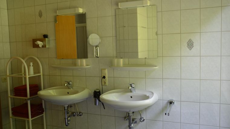 Ein modernes Badezimmer mit zwei Waschbecken und großen Spiegeln. Im Hintergrund sind Handtücher und ein Regal sichtbar.
