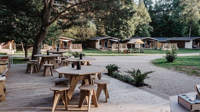 Eine gemütliche Terrasse mit Holztischen und Stühlen, umgeben von Bäumen. Im Hintergrund sind mehrere kleine Holz-Hütten sichtbar.