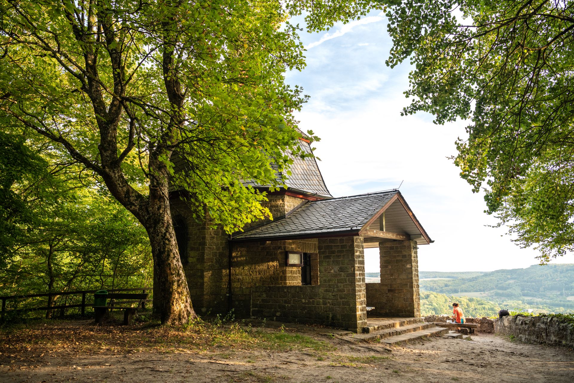 Kleine steinerne Kapelle im Grünen, umgeben von hohen Bäumen, mit Blick über eine weite hügelige Landschaft. Sonnenlicht fällt durch das Blätterdach und betont die ruhige Lage oberhalb des Tals.