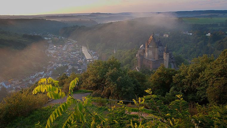 View of the Vianden valley with castle and river, surrounded by green hills.
