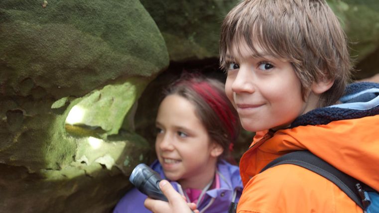 Zwei Kinder erkunden eine Felsformation, während eines das Licht einer Taschenlampe hält. Sie wirken neugierig und begeistert von ihrem Abenteuer.