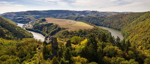 Eine malerische Aussicht auf ein Tal mit sanften Hügeln und einem Fluss. Im Vordergrund sind Überreste einer alten Burg und üppiges Grün zu sehen.