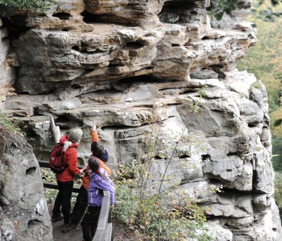 Drei Wanderer auf einem Pfad in der Teufelsschlucht, die beeindruckende Felsformationen betrachten und darauf zeigen., &copy; Felsenland S&uuml;deifel Tourismus GmbH