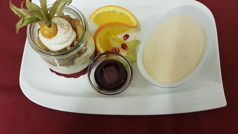 Ein Dessertteller mit einem Glas Süßspeise, dekoriert mit Sahne und einer Beerenfrucht. Dazu gibt es eine Vanillesauce und frische Früchte wie Orange und Kiwi.