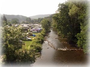 Ein ruhiger Fluss fließt neben einem Campingplatz, umgeben von Bäumen. Im Hintergrund sind Zelte und Wohnmobile sichtbar.