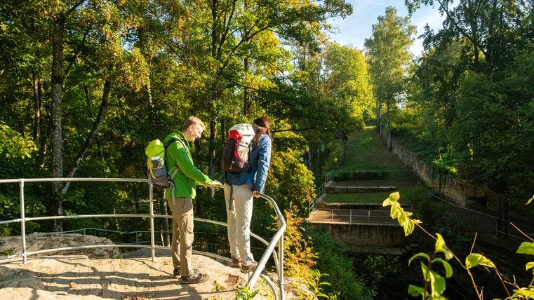 Zwei Wanderer stehen auf einem Aussichtspunkt umgeben von Bäumen. Die Sonne scheint und die Szene vermittelt eine friedliche Naturatmosphäre.