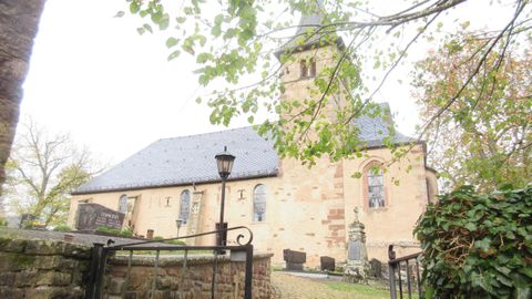 Eine alte Kirche mit einem hohen Turm ist von Bäumen umgeben. Der Himmel ist grau und bewölkt.
