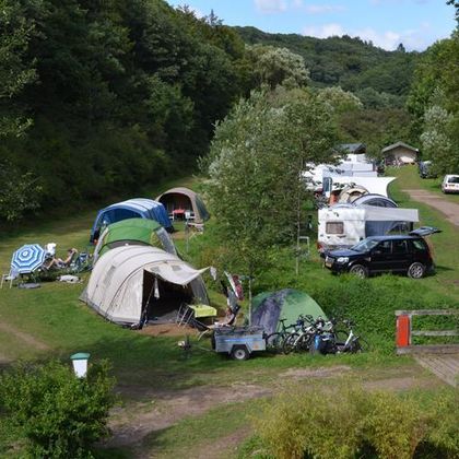 Ein malerischer Campingplatz mit mehreren Zelten und Wohnmobilen in einer grünen Umgebung. Bäume und Sträucher umgeben den Platz, der von einem Weg durchzogen wird.