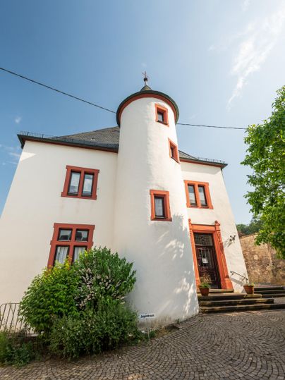 Ein historisches Gebäude mit einer runden Turmstruktur und weißer Fassade. Umgeben von grünem Laub und einer gepflasterten Fläche.