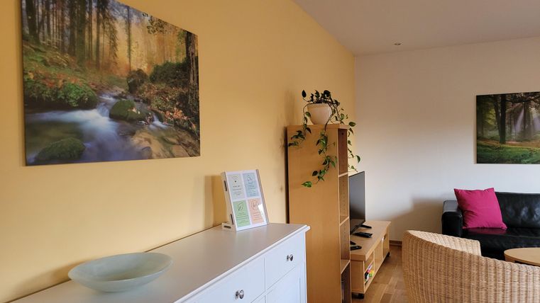 Ein gemütlicher Wohnraum mit einem hellen, gelben Farbton. An den Wänden hängen Bilder von Natur, während ein bequemer Sessel und ein Fernseher den Raum ergänzen.