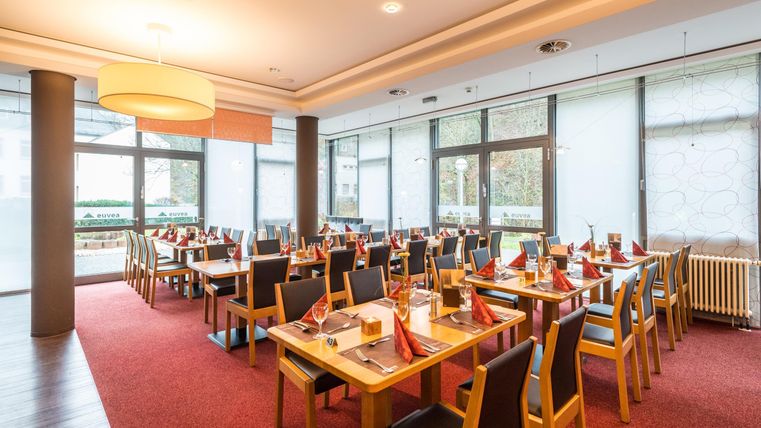 Ein modernes Restaurant mit eleganten Tischen und roten Tischdecken. Große Fenster sorgen für viel Tageslicht und eine angenehme Atmosphäre.