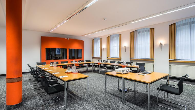 Ein modernes Konferenzzimmer mit mehreren Tischen und Stühlen. Die Wände sind orange und es gibt große Fenster mit Vorhängen.
