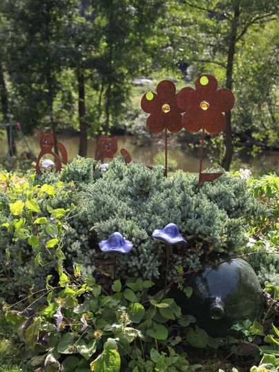 Ein bunter Garten mit grünen Pflanzen und blauen Blumen. Metallische Blumenornamente stehen im Vordergrund.