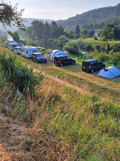 Ein Campingeplatz in der Natur mit mehreren Wohnmobilen und Zelten. Im Hintergrund sind sanfte Hügel und ein klarer Himmel zu sehen.