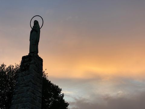 Eine Statue mit einem Heiligenschein steht auf einem hohen Sockel. Der Himmel ist in sanften Farben bei Sonnenuntergang getaucht.
