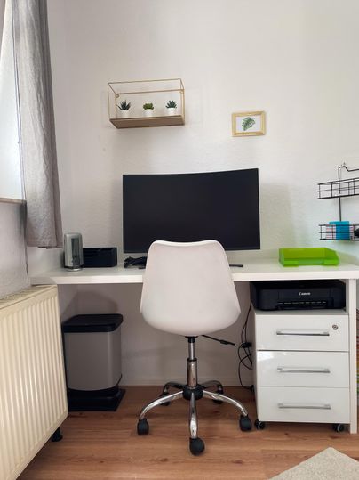 Ein moderner Schreibtisch mit einem Computerbildschirm und einem weißen Bürostuhl. Ein Drucker ist ebenfalls vor Ort.