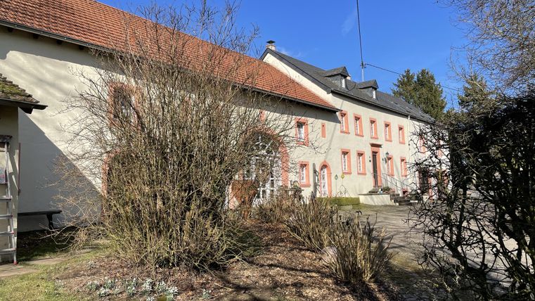 Ein älteres Wohnhaus mit roten Fensterrahmen und einem Garten. Die Umgebung ist grün und sonnig.