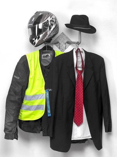 Ein Motorradhelm und eine neonfarbene Jacke hängen neben einem formellen Anzug mit Fliege und Hut. Die Kombination zeigt einen Kontrast zwischen Freizeit- und Abendgarderobe.