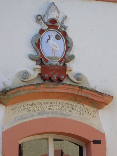Ein Wappen mit einem Storch und einem medaillonartigen Hintergrund ist an einer Gebäudewand angebracht. Unter dem Wappen befindet sich eine Inschrift in geschwungener Schrift.