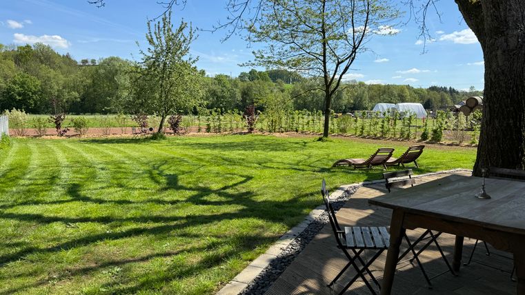 Un beau jardin avec du gazon vert, des arbres et un paysage ensoleillé. Au premier plan se trouvent une table et des chaises, idéales pour se détendre.