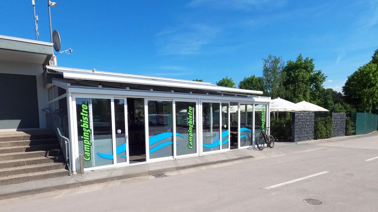 Ein modernes Gebäude mit großen Fenstern und einer hellblauen Fassade. Draußen stehen Sonnenschirme und es ist ein sonniger Tag.