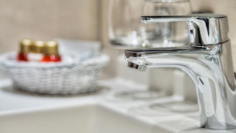 Ein moderner Wasserhahn in einem Badezimmer. Im Hintergrund sind Gläser und ein Korb mit kleinen Flaschen zu sehen.