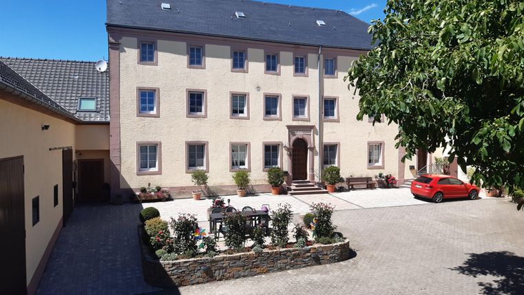 Ein großes, helles Gebäude mit einem geräumigen Innenhof. Im Vordergrund sind Blumenbeete und ein rotes Auto zu sehen.