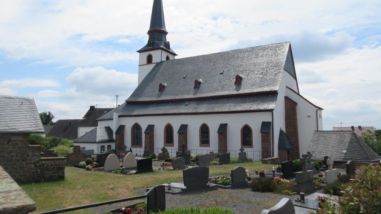 Eine Kirche mit einem spitzen Turm und einem gemütlichen Friedhof im Vordergrund. Der Himmel ist bewölkt und es ist eine ruhige Atmosphäre.