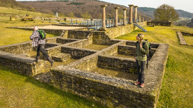 Zwei Personen erkunden die Ruinen der römischen Villa Holsthum mit Säulen und Steinmauern im Hintergrund.