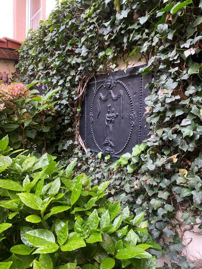 Ein schmiedeeisernes Schild, umgeben von grünen Pflanzen und Efeu. Die Umgebung ist ruhig und naturnah.