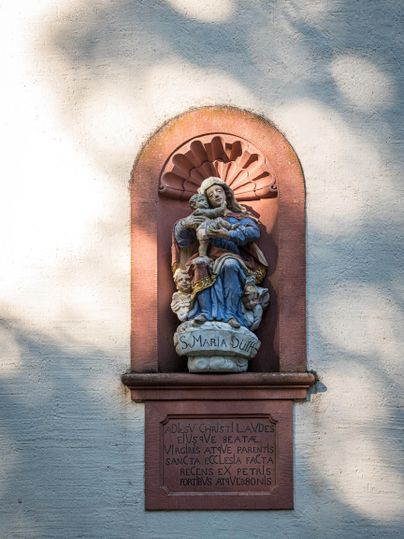 Eine Statue der Maria mit dem Jesukind, die an einer Wand angebracht ist. Sie ist von Licht und Schatten umgeben.