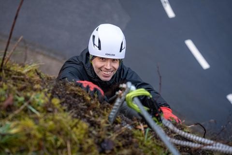 Ein Kletterer mit Helm lächelt, während er an einer steilen Felswand hochklettert. Die Umgebung ist grün und der Weg ist im Hintergrund sichtbar.