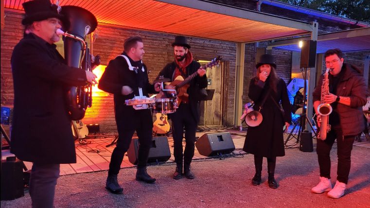 A band is playing live music in a cozy outdoor area. The artists are dressed in black and there is colored light in the background.