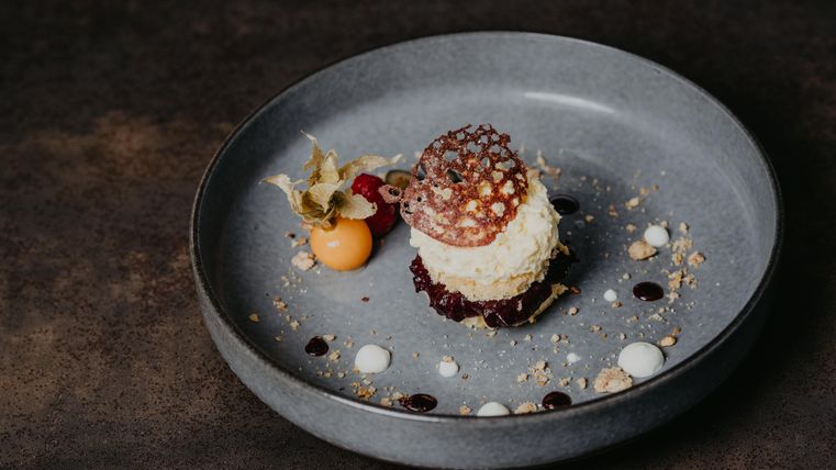 Ein kunstvoll angerichtetes Dessert auf einem grauen Teller. Es besteht aus einer Creme, Früchten und dekorativen Soßen.