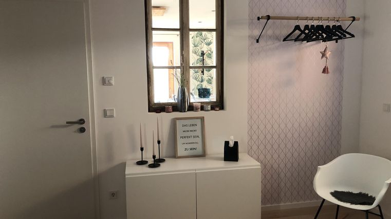 Een lichte kamer met een raam en een wit dressoir. Aan de muur hangt een kledingstang met kledingstukken.