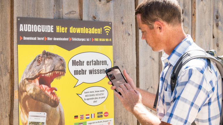 Ein Mann scannt mit seinem Smartphone einen QR-Code auf einem Plakat. Das Plakat enthält Informationen über Audioguides und zeigt ein Bild eines Dinosauriers.