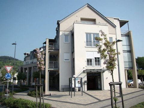 Ein modernes Gebäude mit klaren Linien und großen Fenstern. Vor dem Gebäude sind bepflanzte Flächen und eine kleine Straße sichtbar.