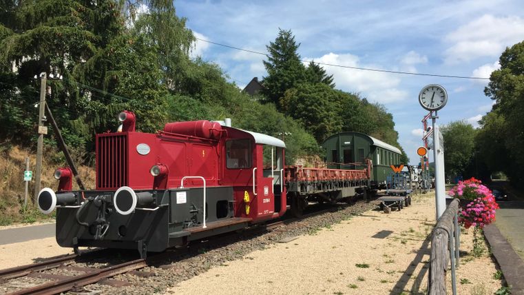 Alter Bahnhof in Pronsfeld mit roter Lokomotive und alten Waggons auf Gleisen, umgeben von Bäumen und Blumen.