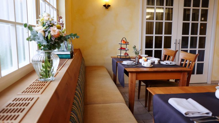 Ein gemütlicher Essbereich mit Holztischen und Stühlen. Auf der Fensterbank steht eine Blumenvase und ein kleiner Dessertständer.